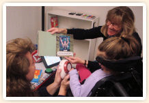 Instructor demonstrates communication devices for speecTwo women showing a girl how to touch a device that reads the text of a book page